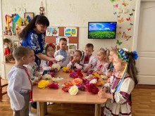 Практикантка педколеджу навчає дошкільнят українським традиціям, створюючи квіти. Діти у вишиванках, навколо українська символіка.