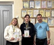 Спортсменки разом з тренером, дівчата тримають подяки, на шиях висять медалі, на задньому плані дошка з подяками.