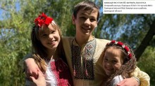Троє усміхнених дітей у вишиванках обіймаються на фоні зелених дерев.