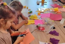 Діти займаються рукоділлям за столом, використовуючи кольоровий папір і ножиці. Атмосфера зосереджена і творча, навколо них розкладені різнокольорові папери рожевого, жовтого та оранжевого кольорів.