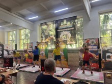 Спортсмени з гирьового спорту. В руках у спортсменів гирі, які вони підіймають однією рукою над головою. Учасники одягнені у спортивний одяг.