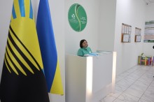 Хол медичного закладу.
Жінка адміністратор стоїть біля стійки приймання громадян.