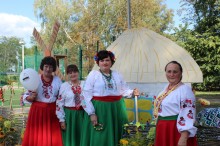 Жінки у національному вбранні, зелені та червоні спідниці, зверху одягнені вишиванки та намисто. Поруч макет української хати