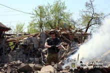 Поліцейський у повній екіпіровці, тримає уламок снаряда. На задньому плані дим та уламки.
