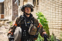 Рятувальник Донеччини. У повній екіпіровці та в броніжелеті. У руках тримає гідравлічний шланг. Обличчя у сажі після пожежі. На задньому плані будинок без вікон.