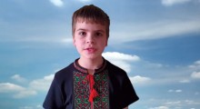 Хлопчик у чорній вишиванці з червоним орнаментом на фоні блакитного неба читає вірш