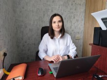 Молода жінка в білій сорочці сидить за робочим столом перед відкритим ноутбуком Dell на фоні сірої текстурованої стіни.