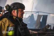 Обличчя рятувальника крупним планом. Чоловік стоїть у розбитій будівлі, все ще в диму. На голові каска з бодікамерою, на бронежилеті прикріплена рація.