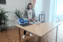 Ведуча стоїть біля столу з ноутбуком, на екрані якого презентація, і VR-окулярами
