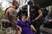 Літня жінка в інвалідному візку, волонтер та співробітники поліції докладають зусиль та заносять її до броньованого автомобіля. Евакуація з прифронтових міст.