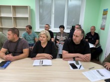 Учасники наради сидять за столом. На столі лежать відкриті блокноти та ручки. Всі уважно слухають.
