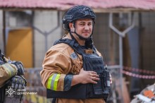 Рятувальник у захисному костюмі та бронежилеті, дивиться у сторону фотографа. На задньому плані понівечені будівлі.