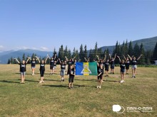 Діти в однакових чорного кольору футболках стоять на галявині, руки підняті вгору, тримають прапор громади. На задньому плані смереки та блакитне небо.