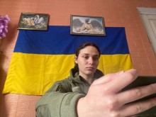 Анастасія Кулікова у військовій формі сидить перед смартфоном на тлі прапора України та картин на стіні.