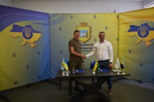 Голови громад тиснуть друг другу руки. Стоять біля столу, на якому лежать документи та стоять прапори громад.