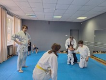 Спортивний зал. Всі спортсмени у білих костюмах. В залі маленький хлопчик, який за допомогою тренера навчається навичкам прийому.