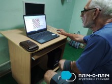 На екрані монітора віртуальна дошка з шахами. Біля монітора сидить чоловік похилого віку в окулярах.