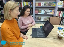 Дві жінки сидять за столом, зайняті роботою на ноутбуці. На задньому плані видно полиці, заповнені книгами. Атмосфера зосереджена і сприятлива для співпраці.