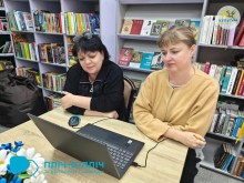 Дві жінки сидять за столом з ноутбуком, зосереджено дивлячись на екран. За ними книжкові полиці заповнені різнокольоровими книгами. Атмосфера навчальна.