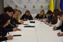 Службовці обладміністрації за столом зосереджено пишуть диктант у залі з прапорами України та символікою Донецької області.