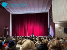 Глядацька зала театру, повна людей; на сцені перед закритою червоною завісою стоїть промовець.