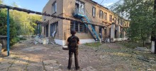 Правоохоронець стоїть на фоні пошкоджено\ обстрілом двоповерхової будівлі.