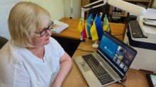 Учасниця вікторини сидить біля ноутбука, поруч стоять прапорці.
На екрані монітора заставка проєкту «Пліч-о-пліч: згуртовані громади» та інші учасники.