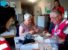 Троє представників Благодійної організації та жінка похилого віку сидять за столом, на столі лежать медичні прилади. Жінка тримає у руках таблетки та щось розповідає