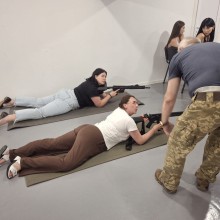 Дві жінки уважно слухають інструктора, який пояснює основи стрільби з автомата лежачи.