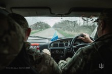 Салон автомобіля. Руки військового тримають кермо, поруч сидить побратим. Спереду дорога та посадка.