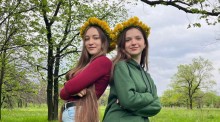 Дві дівчини підліткового віку у вінках із кульбаби стоять спинами одна до одної на зеленій галявині. Обидві посміхаються.