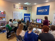 Жінки уважно слухають розповідь учасниці зустрічі, яка стоїть біля екрану монітора. На стінах висять прапори організаторів Smarta 
«МиРазом» Центр підтримки ВПО Краматорського району