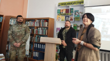 Ведуча виступає перед аудиторією в бібліотеці, поруч із нею стоять два чоловіки у військовій формі