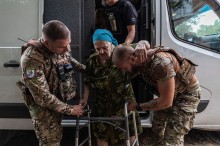 Співробітники поліції допомагають літній жінці, одного з поліцейських жінка обіймає рукою за шию, іншою рукою тримає ходунки.