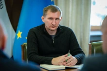 Учасник робочої наради. Сидить за столом, тримає ручку, на столі лежить відкритий щоденник