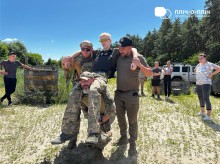 Інструктори та учасники практичного курсу з тактичної медицини тримають учасника курсу за руки, третій інструктор підтримує його тазову частину своєю спиною, сидячі на колінах