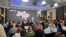 Великий зал, наповнений людьми. На сцені стоїть жінка у світло-синьому костюмі та звертається до аудиторії. За нею на великому екрані відображено карту України з назвами областей. Зліва від карти на екрані також видно її зменшене зображення.