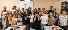 A group of smiling children and teens pose indoors in front of a banner. They are gathered around tables with art supplies, exuding joy and creativity.