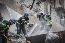 Пожежники в захисному спорядженні прибирають уламки з пошкодженої будівлі. Місце події заповнене пилом і уламками, що створює атмосферу терміновості та стійкості.