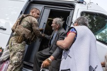 Поліцейський допомагає чоловіку похилого віку сісти в броньований автомобіль, поруч стоїть жінка похилого віку.