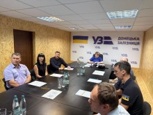 Представники регіональної філії "Донецька залізниця" АТ "Укрзалізниця" сидять за великим столом у конференц-залі і є учасниками онлайн сесії в межах ярмарку вакансій. На задньому плані розміщений банер з логотипом "Укрзалізниця" та написом "Донецька залізниця".