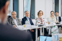 Четверо учасників круглого столу уважно слухають колегу, серед них жінка у білій вишиванці з червоним орнаментом.