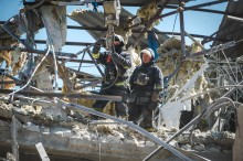 Двоє рятувальників у захисному спорядженні пробираються крізь уламки зруйнованої будівлі, оточені скрученим металом та ізоляційним матеріалом, під чистим небом.