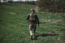 Захисник України. Фото зі спини. Йде по зеленій траві. Одягнений у повній екіпіровці.