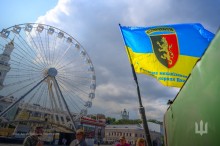 Прапор сухопутних військ, 24 ОМБр імені короля Данила майорить на фоні атракціону колеса огляду. Дія відбувається на Контрактовій площі, місто Київ.