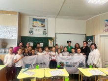Діти у шкільному класі, тримають прапори громад. Позаду шкільна дошка, попереду - парти жовтого кольору.