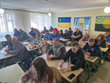Представники управління фізичної культури та спорту сидять за партою в кімнаті з українськими прапорами та пишуть диктант.