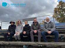 Група з п'яти осіб сидить на відкритих трибунах під хмарним небом. Вони одягнені в куртки, що свідчить про прохолодну погоду. Настрій здається спокійним і невимушеним.