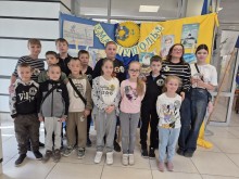 Група дітей і двоє дорослих стоять, посміхаючись, на тлі барвистого фону з малюнками та текстом кирилицею. Сцена яскрава і весела.