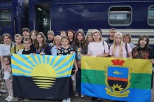 Діти та дорослі стоять на пероні вокзалу. Позаду них вагон потяга синього кольору. На передньому плані діти тримають прапори громади та Донеччини.